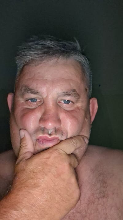 A close-up, waist-up, natural light portrait features a Caucasian man with short, graying hair. He is looking directly at the camera, with his right hand resting on his chin and mouth area. The man has blue eyes and a slightly pursed mouth. He is shirtless, and his skin appears flushed. The background is dark and out of focus, with a hint of a green wall visible on the right side of the image. The setting is indoors, and no specific location within Voinova, Moldova, is discernible from the image.