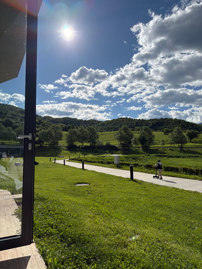 A slightly rotated, outdoor scene shows a bright sunny day with a clear, blue sky and fluffy white clouds over a grassy, green landscape with trees and a hill in the distance. The sun is bright, creating a lens flare in the upper left. A paved path runs along the edge of the grass, and a person on a scooter is visible on the path. There are short, black posts lining the path. The foreground shows part of a building, possibly a window frame or a door frame, with a handle. The scene appears to be in Chkalov, Armenia, judging by the landscape and architecture.