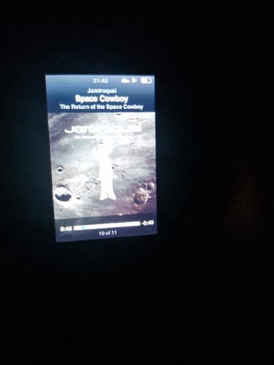 The image shows a mobile phone screen displaying the song "Space Cowboy" by Jamiroquai from the album "The Return of the Space Cowboy." The phone's display is black and the screen is lit up, showing the album art, song title, artist name, album name, time elapsed (0:46), total song time (5:40), and the track number (10 of 11). The album art is a black and white image of a person standing on a rocky surface with craters in the background. The phone is in a dark room with a black background. The coordinates provided place the phone in the town of Hertford, East Hertfordshire, England.