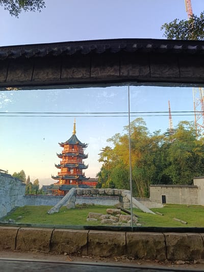 This image captures a scene at the Surabaya Zoo in Durin Serugun, Indonesia, as evidenced by the distinct architectural style of the panda enclosure and the prominent pagoda structure visible in the background, which is a well-known feature of the zoo. The foreground shows a barrier made of rough-hewn, segmented stone blocks, likely a part of the enclosure's viewing area for visitors. Above this barrier, a large glass pane reflects the sky and trees, but also allows a clear view into the enclosure.
Inside the enclosure, a lush green lawn is dotted with various rocks and logs, designed to mimic a natural habitat. A concrete structure with a dark door is visible on the right side of the enclosure, possibly a shelter or a service building. Bamboo stalks and other dense foliage form a naturalistic backdrop to the enclosure, with the iconic multi-tiered pagoda with its dark, curved roofs and golden finial rising majestically behind it. The pagoda, painted in vibrant red and green, is a clear architectural landmark. In the distance, two communication towers, one red and white and another taller, are visible against the clear blue sky, indicating the presence of urban infrastructure near the zoo. The lighting suggests it is either early morning or late afternoon, with soft sunlight illuminating the scene and casting gentle shadows. No people or animals are clearly discernible within the enclosure in this particular shot.