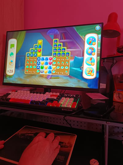 The image captures a first-person perspective of someone playing a mobile game on a computer monitor. The game appears to be a match-three puzzle game, with colorful gems and game pieces arranged in a pyramid-like structure on the screen. On the left side of the screen, there are game statistics showing "44", "20", "19", and "$25". To the right of the game board, a vertical menu displays icons for game power-ups: a hammer, a watering can, and a fan.

The monitor is an AOC brand. Below the monitor, a white and red mechanical keyboard with RGB lighting is visible, displaying the brand "XGLOWS". A mousepad with a dark image is also present, with a hand resting on it, suggesting active gameplay.

The setting is likely a home or office environment, illuminated by a red lamp that casts a reddish hue over the scene, particularly on the walls and the user's hand. A red desk lamp is positioned to the right of the monitor. In the background, shelves with books and a blue sofa can be seen, indicating an indoor setting, possibly a living room or a dedicated gaming space. A small analog clock with an orange face is also visible on a shelf, showing a time between 10 and 11. The overall atmosphere is dimly lit, with the dominant colors being red and pink from the lighting, and the bright, vibrant colors of the game on the screen. The image does not provide information about the time of day or weather conditions.