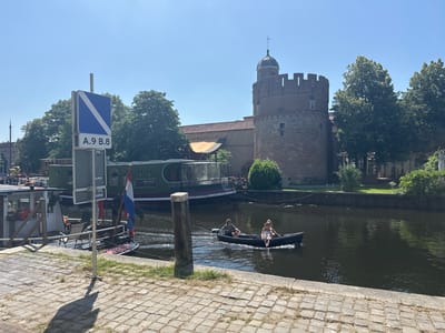 This is a horizontal, eye-level outdoor photograph taken on a sunny day. The scene depicts a canal in what appears to be an urban setting with historical architecture. In the foreground, a cobblestone walkway lines the edge of the canal.

On the canal, a dark-colored canoe is being paddled by two individuals. A man in a grey and white striped shirt is in the front, and a woman in a white tank top and light-colored shorts is in the back. They are both looking forward as they move through the water.

To the left of the canoe, a larger, dark green boat is docked. Part of this boat is visible, showing windows and what appears to be a covered seating area. A Dutch flag is prominently displayed on a smaller boat docked behind the green one. A tall sign with a blue and white directional symbol and the text "A.9 B.8" is also visible on the left side of the frame, near the docked boats.

In the background, a prominent, round, brick tower with battlements and a domed roof stands out against a clear blue sky. Adjacent to the tower is a building with red-tiled roofing and more brick structures. Lush green trees line the canal banks and surround the buildings.

The sunlight suggests it is daytime, likely midday or early afternoon, given the bright conditions and the shadows cast. The overall atmosphere is serene and leisurely, with the people in the canoe enjoying a calm day on the water. The presence of the Dutch flag and the canal setting are strong indicators of a location in the Netherlands, specifically Zwolle. Text visible on the boats includes "BUKO", "PRODUCHTHUIS", and "WOLFS". There is also a sign on the boat in the foreground with the Dutch word "BETREDEN" which means "entered" or "entered".