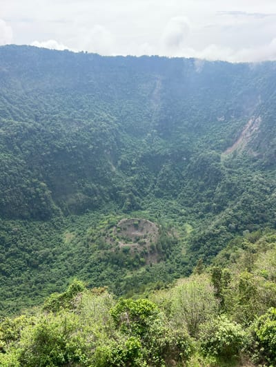 The image depicts a mountainside covered in dense green vegetation, possibly located in or near Santa Tecla, El Salvador. The left side of the image shows a steep, rocky cliff face partially obscured by mist or haze. The vegetation consists of trees and smaller plants, creating a varied texture. In the middle of the image, there is a circular clearing, possibly a landslide or a deforested area. The right side features a steeper slope with slightly different vegetation, possibly closer to the camera, including branches and leaves of trees.