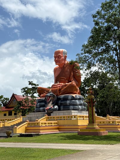 This is a full, eye-level, outdoor, vertical, medium shot of a large, orange-colored statue of a seated Buddhist monk in front of a bright blue sky with scattered white clouds. The monk statue is the dominant subject, positioned centrally and slightly to the left of frame. It is seated in a cross-legged meditative pose on what appears to be a coiled, black-and-white serpent sculpture. The monk's face is wrinkled, and he has a serious expression. He is wearing saffron-colored robes.
To the left of the statue, a yellow and red temple building with a red tiled roof is visible. Behind the statue and to the right, lush green trees with dense foliage are present. In the foreground, a well-maintained green lawn is visible, with a concrete pathway cutting through it. A brightly colored, ornate yellow and red platform with staircases and railings surrounds the base of the statue. A decorative, red and yellow pillar is positioned to the right of the statue's base. The overall setting appears to be a religious or cultural site in an outdoor environment. This statue is located in the city of Na Nuea, Thailand.