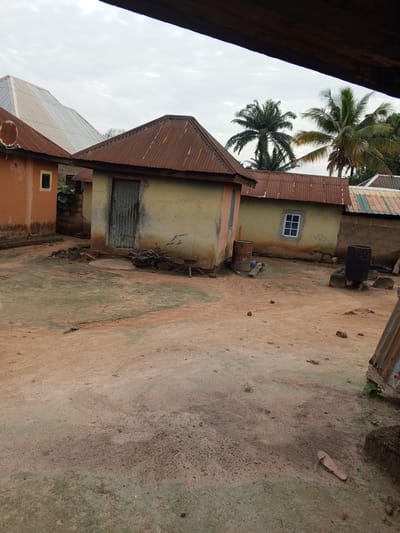 The media file depicts a scene in Gindin Dutse, Nigeria, featuring several small, weathered buildings with corrugated tin roofs. The foreground is a dusty, dirt yard with scattered stones and patches of sparse vegetation.
In the midground, a light yellow building with a rusty tin roof has a closed, metal door. To the left of this building, an orange-colored structure with a smaller, dark rectangular window is visible. Further to the right, another building with a similar tin roof has a white-framed window with multiple panes. Two large, rusted metal barrels stand outside the yellow building, one near the door and another further to the right. A small white chicken is near the barrels on the right. In the background, palm trees are visible, suggesting a tropical or sub-tropical climate.
The sky is overcast and appears to be either early morning or late afternoon due to the diffused light. There is no overt activity taking place; the scene appears still and quiet. Notable details include the worn state of the buildings, the rustic nature of the objects, and the generally dry, dusty environment. There is some faint text visible on the yellow building to the right of the door, but it is illegible. The overall impression is one of a simple, rural settlement.
