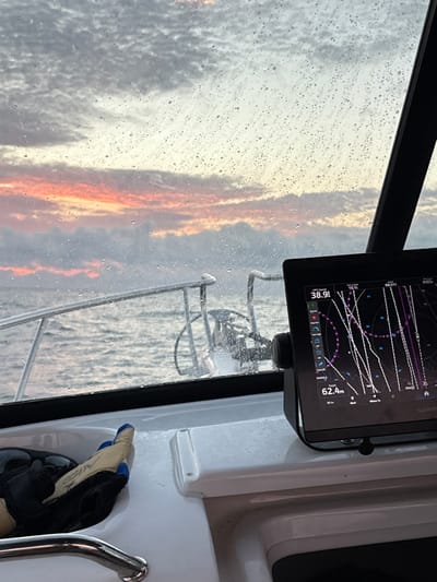 The view from inside a boat's cabin shows a rainy window with a vibrant orange and grey sunset. A Garmin GPS screen displays a navigation map with multiple routes. Outside, a stainless steel railing is visible. Inside the cabin, a pair of gloves rests near a white panel. The scene suggests a journey underway, possibly during inclement weather, with the boat's systems actively guiding the vessel.