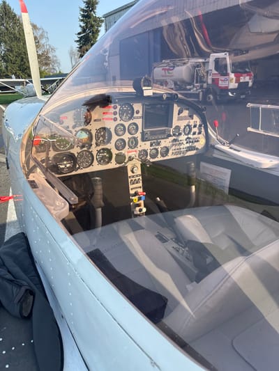 The image presents an exterior view of a light-grey fixed-wing aircraft positioned on an asphalt or tarmac surface at an airfield in Toussus-le-Noble, France. The perspective is from the aircraft's left side, looking into the cockpit through its closed, transparent canopy. The cockpit interior reveals a detailed instrument panel comprising multiple circular analog gauges, a central rectangular digital display unit, and a vertical column of assorted switches and circuit breakers on the right side. A control stick is visible below the instrument panel. Two light-grey upholstered seats are present within the cockpit. On the exterior of the aircraft's fuselage, a black jacket or garment is draped over the body near the cockpit opening. A red warning flag or cover is partially visible on the far left edge of the frame. In the background, beyond the main aircraft, are the tail section and propeller of a second white aircraft, a large metal-clad hangar building, and a white and red TotalEnergies AVGAS fuel truck. Green trees line the background under a clear, bright blue sky. No individuals are visible.