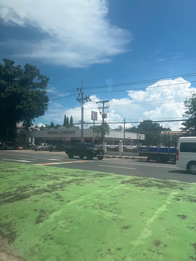 The image shows a street scene in Ban Tha Chat Chai, Thailand on a sunny day with scattered clouds. A bright green surface, likely a bike lane or path, occupies the foreground, with dark, irregular markings suggesting wear and tear. Beyond this, a paved road runs alongside it.
Several vehicles are visible on the road, including a dark-colored SUV parked on the right side of the road and a silver pickup truck further down. A white van is also present, as well as a dark-colored open-bed truck. In the background, a 7-Eleven convenience store is clearly identifiable by its distinctive red and green signage and building design. Electrical poles with multiple wires stretch across the sky, common infrastructure in the area. Trees, some with dense foliage, frame the scene, adding to the tropical feel. The sky is a vibrant blue, with fluffy white clouds indicating fair weather. The overall impression is of a typical daytime scene in a Thai town, likely during daylight hours given the bright sunshine. No people are explicitly visible in the frame. The image is oriented vertically, with the sky and power lines dominating the left side and the road and buildings on the right.