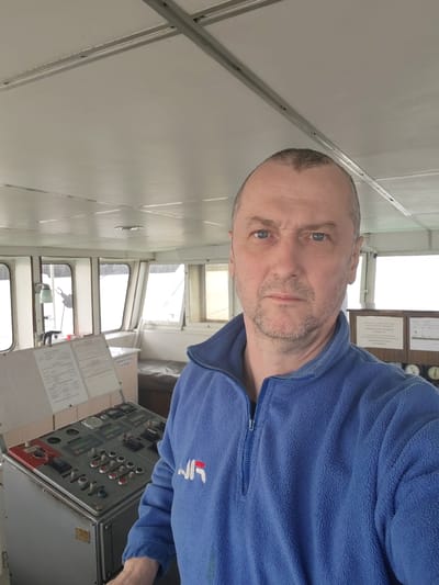 A middle-aged man stands in the control room of a vessel, possibly a boat or a ship. The control room has numerous windows offering a bright view of the outside. A control panel with buttons, switches, and digital displays stands to the left of the man. Behind him, a bulletin board is visible with papers and gauges. The man is wearing a blue fleece jacket. The setting appears to be an interior space of a watercraft, but there is no confirmed location of Khoroshevo, Russia.