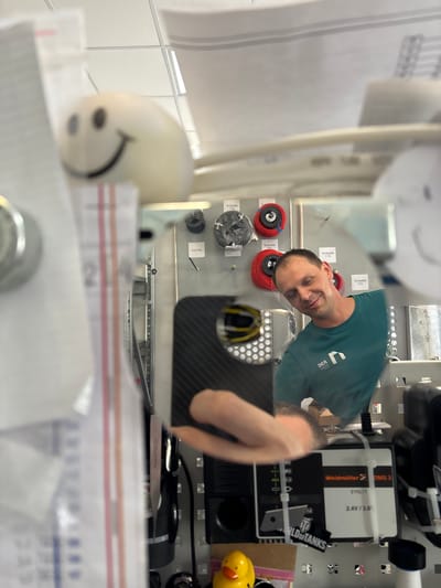 This is a portrait-style, slightly blurry, and artificially lit photograph taken at eye level. The image is oriented sideways. In the foreground, there's a blurry white object with a smiley face drawn on it. Behind this, a man in a teal t-shirt with a white logo is visible, smiling with his eyes closed. He appears to be in a workshop or lab setting, with various objects and equipment in the background. These include spools of wire, electronic components, and what looks like a carbon fiber surface. Text visible in the image includes "Weidmüller DMS 3," "EYOL11," "2.4V/3.6," and "RLDOFTANKS." The background also shows a white tiled wall and shelves with more items. The overall impression is a candid, informal shot of someone in their workspace.