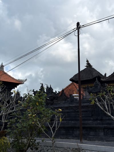 The image shows a street view in Susut, Indonesia, under a cloudy sky. In the foreground, a dark stone wall, typical of Balinese architecture, forms the boundary of a property. Several trees with delicate branches and sparse foliage, some bearing small yellow flowers, are visible in front of the wall. Behind the wall, elements of traditional Balinese buildings can be seen, including terracotta tiled roofs and thatched roofs topped with ornate spires. A prominent utility pole with multiple dark wires stretches across the upper portion of the frame, obscuring some of the sky and architectural details. The overall impression is one of a rural or semi-rural setting in Indonesia, likely a temple or a traditional residence, with everyday infrastructure elements present.