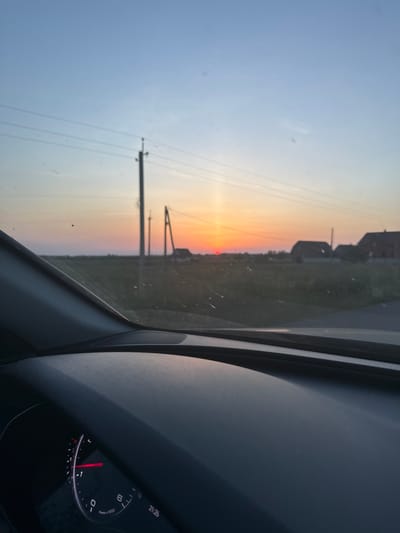 The image depicts a sunset viewed from inside a car. The sun, a small red disc, is low on the horizon, casting a warm orange and yellow glow across the sky. Silhouetted against the vibrant sky are power lines and a few buildings, suggesting a rural or semi-rural setting. The foreground is dominated by the interior of the car, with a dashboard and a speaker visible on the right side. The dashboard shows a speedometer with a red needle and some visible text, including "21:25," indicating the time is 9:25 PM. The overall mood is serene and peaceful, capturing the beauty of a sunset in Dovhe, Ukraine. The weather appears to be clear, with no clouds obscuring the sunset. The image is oriented vertically, with the sunset appearing on the left side and the car interior on the right.
