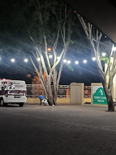 The image captures an outdoor scene at night, illuminated by string lights hanging between several trees with light-colored trunks. The ground is paved with dark-colored blocks or cobblestones.
On the left side, a white ambulance is parked. The side of the ambulance features an image of a man, likely a political figure, and the text "PRABOWO" above "AMBULANCE". A partial Indonesian license plate, "B 2018 GO", is visible on the rear.
In the mid-ground, behind the ambulance and trees, there is a low fence or wall with ornate white metalwork on top, separating the paved area from a dimly lit background structure. Near the base of one of the trees, a blue tarp and a white sack are resting on the ground.
On the right side of the image, behind another tree, a green sign with white text and a white pictogram is visible. The text reads "TEMPAT WUDH" (likely "Tempat Wudhu" for ablution place) and "WANITA" (Women), indicating a facility for female ablution.
The overall setting appears to be a public or semi-public area, possibly near a mosque or community center, given the sign for ablution. The string lights create a festive or welcoming atmosphere. The time of day is clearly night, with no specific weather conditions apparent. The branding on the ambulance suggests a political campaign or affiliated service. The Indonesian text on both the ambulance and the sign confirms the location context of Indonesia. No people are visible, and no active event is taking place; the scene is static.