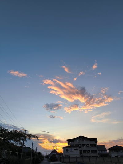 The image depicts a sunset over a residential area in Berehove rural council, Ukraine. The sky is a gradient of blue and orange, with scattered clouds illuminated by the setting sun. In the foreground, a paved road curves through a village lined with houses and trees. Utility poles and wires are visible along the roadside. The architecture of the houses suggests a mix of modern and traditional styles. The overall scene evokes a sense of tranquility and rural life.