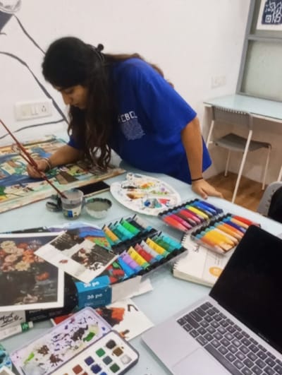 In Aktash, Uzbekistan, a female individual with long, dark hair, wearing a blue t-shirt, leans forward at a light-colored table, actively engaged in painting. She holds a paintbrush in her right hand, applying pigment to an unfinished artwork depicting a multi-colored scene. The table is densely covered with various art supplies: multiple paintbrushes, a glass with water, an oval-shaped artist's palette displaying mixed paints, several organized sets of colorful paint tubes in plastic trays, a rectangular watercolor pan set, loose reference images (including one of flowers), a smartphone, a notebook, and a partially visible silver laptop on the right. The background consists of a plain white wall with electrical outlets, a partial black outline mural, and a secondary light-colored desk with a gray chair.