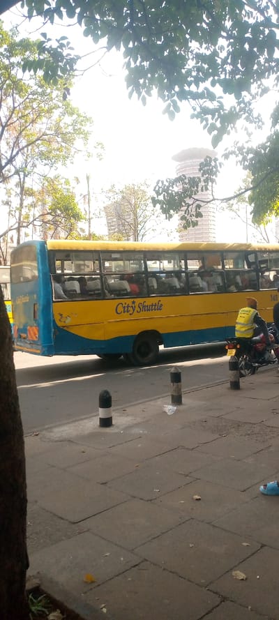 A yellow and blue "City Shuttle" bus drives along a city street, filled with passengers. A motorcyclist with a yellow vest pauses nearby. Black and white bollards line the curb, and a piece of trash sits on the sidewalk. In the background, a tall building, likely a landmark, rises behind trees. The scene is bathed in bright daylight, with trees casting shadows in the foreground.