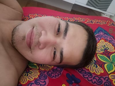 A close-up, high-angle, eye-level shot shows a young adult male lying on his back on a red fabric with colorful floral patterns. The male is positioned on his left side, with his head tilted towards the camera. His face is the primary focus, with his right eye, nose, and mouth visible. He has dark eyebrows and a light beard growth on his chin and upper lip. His skin is fair. The background consists of a wooden surface and a glimpse of a white furniture piece. There are no discernible features that place this image specifically within the city of Zhanaozen, Kazakhstan.