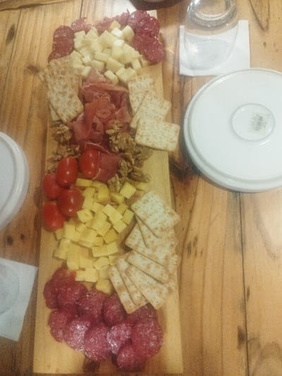 A wooden board holds an array of food items. Slices of salami bookend the arrangement. Between them are cubes of cheese, stacks of crackers, prosciutto, walnuts, and cherry tomatoes. A glass of water and stacked white plates sit beside the board. The surface below is a wooden table.