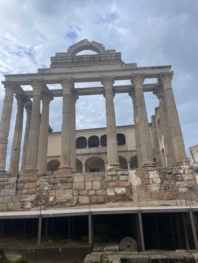 This image captures the ancient Roman ruins of a temple, identified as the Temple of Diana in Mérida, Spain. The photograph is taken from a low-angle perspective, looking up at the monumental stone structures against an overcast sky.
The primary subjects are a series of tall, weathered stone columns, likely of the Corinthian order, distinguished by their fluted shafts and intricately carved capitals. These columns support a partially preserved entablature, which includes a fragmented pediment. A prominent architectural feature is a distinct, half-arch element on the upper left side of the entablature.
To the right of the main column structure, a section of an older, robust stone wall or foundation is visible, constructed from irregularly shaped, interlocking blocks. Beyond this wall, an excavated area appears dark and shadowy, containing various stone fragments and a large, circular stone object resembling a millstone or similar ancient implement.
In the background, positioned behind the ancient temple remains, is a more modern, light-colored building. This structure features multiple arched windows and openings on its upper levels, suggesting it may be a later construction built into or adjacent to the historical site.
The sky is largely covered with light and dark grey clouds, indicating an overcast daytime scene, though some lighter areas suggest patches of less dense cloud cover. No people are visible, and no specific activity is taking place within the frame. The scene evokes a sense of historical depth and architectural endurance. There is no visible text in the image.