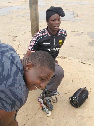 This media file shows two young men outdoors, possibly in Pankshin, Nigeria. One man, wearing a black beret and a black and red athletic shirt with white trim, is lying down with rollerblades on his feet. His shirt has the brand name "UMBRO" visible, along with a yellow crest. He is looking upwards and smiling. The second man, in the foreground, is wearing a grey camouflage t-shirt and is also smiling and looking away from the camera. He appears to be sitting or lying down, facing towards the man with the rollerblades.
In the background, a black portable speaker with orange accents rests on a textured concrete surface. The concrete appears weathered and cracked, with some scattered small, dark spots. A vertical wooden pole runs along the left side of the frame. The lighting suggests it is daytime, with soft shadows, indicating possibly late afternoon or early morning. The overall mood is relaxed and cheerful, with both individuals exhibiting smiles.