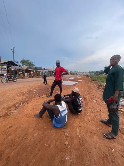 In Osu, Nigeria, on a dusty, red-dirt road under a cloudy sky, several men are gathered. To the right, a man in a green dashiki stands with his right hand holding a red smartphone and his left hand holding a black plastic bag. He is looking off to the left. In the foreground, three men are seated on the ground. One man, wearing a blue and white tank top, is facing away from the camera, seated with his legs extended. Next to him, a man in a black t-shirt with a graphic design and a light-colored baseball cap is crouched, looking down. Partially visible behind him is another man in a black shirt. In the background, a motorcycle is speeding down the road, blurring into motion. The road itself is uneven and appears to be undergoing some form of construction or repair, with muddy puddles and debris scattered around. Buildings with awnings and stalls line the left side of the road, indicating a market or roadside shops. Further down the road, trucks and other vehicles are visible, along with more distant buildings and vegetation.