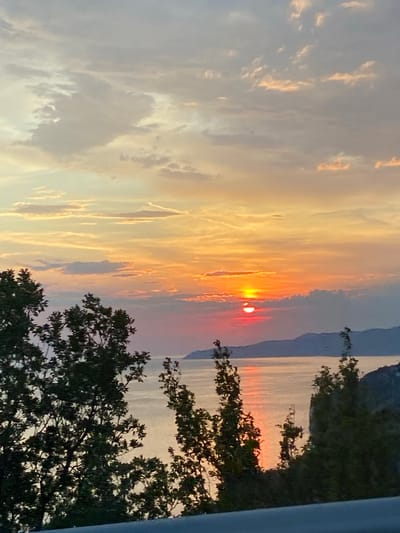 The image depicts a sunset over the sea, viewed from a hillside in Gornji Katun, Montenegro. In the foreground, dark green trees with dense foliage frame the scene on the left and right. The sky is filled with clouds painted in hues of orange, yellow, and pink, with the setting sun a vibrant orb of light near the horizon. A sliver of the sun is visible above a cloud bank, casting a reddish glow. The sea below reflects the colors of the sunset, creating a shimmering surface. In the distance, the silhouette of mountains can be seen against the colorful sky. The overall impression is one of a tranquil and picturesque coastal landscape at dusk.