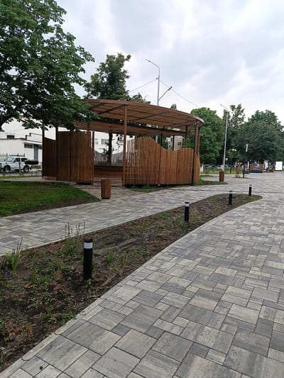 The image depicts a public park in Gubkin, Russia, with a curved paved walkway in the foreground. The walkway is made of interlocking grey pavers with a textured surface. Along the side of the walkway, there are beds of soil with sparse vegetation and small, black bollard-style lights with white tops.

In the middle ground, a contemporary wooden amphitheater or gazebo structure is visible. It features a slatted wooden roof supported by dark posts and a curved, slatted wooden wall enclosing the seating area. A wooden trash receptacle is situated near the entrance to this structure.

Further in the background, mature green trees line the perimeter of the park. Behind the trees, a white building with windows is partially visible. A white vintage car is parked on a paved area to the left of the structure. Streetlights and other park amenities, such as benches and trash receptacles, can be seen along a pathway that recedes into the distance. The sky is overcast with grey clouds, suggesting a cloudy day. The scene appears to be an outdoor public space designed for recreation and relaxation.