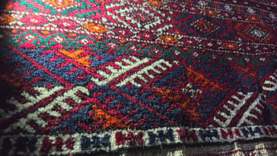 The image shows a close-up view of a richly patterned carpet, likely a Tekke Turkmen rug, with a complex geometric design. The dominant colors are deep red, dark blue, cream, and orange, interspersed with accents of green. The texture appears to be that of woven wool, with a slightly raised pile. The lighting creates dramatic shadows and highlights, emphasizing the texture and patterns of the rug. There is no visible indication of the specific location within Kaka, Turkmenistan, beyond the implication that this is a representation of local craftsmanship or artistry. The scene is static and focused solely on the rug itself.