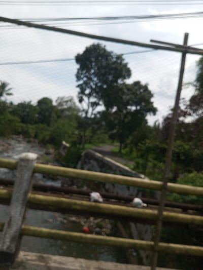 The image depicts an outdoor scene in Namthoung, Laos, captured from an elevated position, likely a bridge or similar structure. The foreground is dominated by elements of a railing system: a light-colored concrete pillar on the left, several horizontal yellowish-brown bars, and a thin, dark vertical pole on the right. Multiple dark horizontal lines, consistent with power or communication cables, span the upper portion of the frame against an overcast sky. A subtle grid-like pattern, possibly a net or screen, is visible across the upper and mid sections of the image.
Below the railing, a body of water, likely a river or stream, flows from left to right, with visible rocks and debris along its banks and within the water. Several small, light-colored objects, resembling discarded plastic bags, are visible resting on the lower horizontal bars of the railing. A distinct red object is partially submerged or located on the riverbed.
In the midground, an arched concrete structure, appearing as a weathered bridge or culvert, spans the water. Beyond this structure, a dark, unpaved or asphalt road curves slightly to the right, disappearing into a densely vegetated area. The background consists of numerous deciduous and evergreen trees, forming a thick canopy of dark green foliage. The sky is largely overcast, displaying light grey cloud cover. No individuals are discernible within the frame, and no specific actions or interactions are depicted. The environment appears rural or semi-rural.