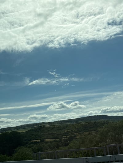 The image displays a wide-angle view of a hilly, rural landscape under a partly cloudy sky. The foreground is dominated by a grey metal railing, likely part of a bridge or overpass, suggesting the viewpoint is elevated. Beyond the railing, dense green trees and shrubs cover the lower slopes of rolling hills. Further back, the hills are less densely vegetated, revealing patches of lighter green fields and darker, more rugged terrain. The sky is a bright blue, streaked with various types of clouds, from wispy cirrus formations to larger, cumulus clouds. There are no people or animals visible in the image. The scene captures a natural, somewhat untamed environment. The provided metadata indicates the location is Blajievo, Bulgaria.