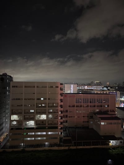 This image captures a nighttime view of an urban landscape in Pasay City, Philippines. The foreground features a dark, overgrown grassy area. Behind this, several multi-story buildings are visible, illuminated by interior and exterior lights.
The prominent building on the left is a tall, white structure with numerous windows and visible air conditioning units. To its right, a slightly shorter, lighter-colored building displays the signage "K LINE" in red letters. This building also has many windows, some of which are illuminated, and air conditioning units. Further to the right, another building exhibits a blue sign that reads "abenson". In the distance, a cityscape with taller buildings and lights can be seen, indicating a dense urban environment. The sky above is dark and cloudy, with some patches of lighter clouds catching the light from the city below. The overall impression is one of a busy city at night, with commercial and residential buildings contributing to the illuminated skyline. No people are directly visible in the image, and no specific actions are depicted beyond the general illumination of the structures.