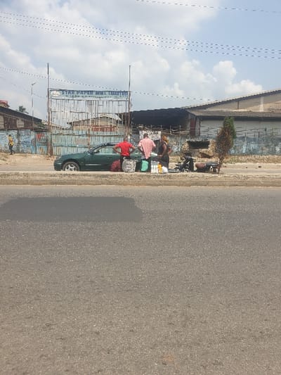 Two men are near a green car parked beside a road. One wears a pink shirt and the other a red shirt. They appear to be selling items on the street. The road surface is gray asphalt. A building with a rusted metal structure and a sign is in the background. Above the building is a cloudy sky with power lines visible. A small, dead tree stands near the edge of the road behind the men.