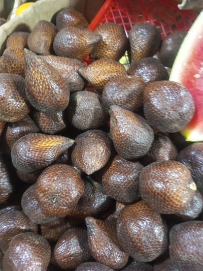 The image displays a close-up, high-angle view of a large, densely packed pile of Salak (snake fruit) in Lê Thanh Nghị Ward, Vietnam. Dozens of individual fruits, characterized by their distinct reddish-brown to dark brown, glossy, and heavily scaled husks, are clustered together, occupying the majority of the frame. Each fruit is typically ovoid to conical, approximately 5-8 cm in length, with the scaly texture resembling reptile skin.
In the upper left quadrant, the edge of a light brown, possibly cardboard or woven, container is visible, holding some of the fruit. Towards the upper right and right side, a segment of a bright red, perforated plastic crate is partially visible, also containing Salak fruit. A thick, triangular slice of ripe watermelon, identifiable by its vibrant red flesh, scattered small black seeds, and a thin green rind, is positioned along the far right edge of the frame, suggesting the broader context of a fruit display. No human figures or direct interactions are visible; the scene is static, focused entirely on the arrangement of produce.