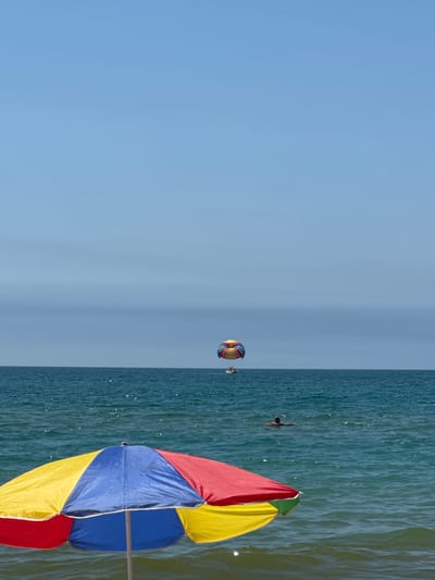 The image depicts a sunny day at the beach in Vityazevo, Russia. In the foreground, a brightly colored beach umbrella with red, yellow, and blue panels stands open on the water's edge. The sea is a deep turquoise, with gentle waves. In the mid-ground, a person is swimming in the water. Further out, a parasail is visible in the sky, attached to a boat. The sky is a clear, light blue.