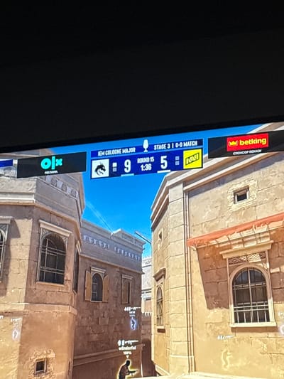 The image shows a screen displaying a video game, specifically Counter-Strike: Global Offensive (CS:GO) during the IEM Cologne Major tournament. The screen displays game information, including the tournament name "IEM COLOGNE MAJOR", the current stage "STAGE 3", the match score "0-0 MATCH", the round number "ROUND 15", and the time remaining "1:36". Team logos and scores are also visible, with one team having a score of "9" and the other "5". Advertising banners for "OLX" and "betking" are present on the screen. The background of the screen shows a virtual environment that appears to be a desert-like city map within the game, with buildings, windows, and a bright blue sky. The image is a photograph of a screen, taken from an angle, with some glare. There is no information in the image that relates to Gothenburg, Sweden.