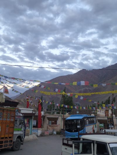 In Alchi, India, a light blue bus is parked on a street in front of buildings. Prayer flags in various colors are strung across the street, and the sky is overcast. A large orange truck is parked to the left of the bus. In the background, a mountain range rises. The street appears to be a commercial area, with shops and signs visible.
