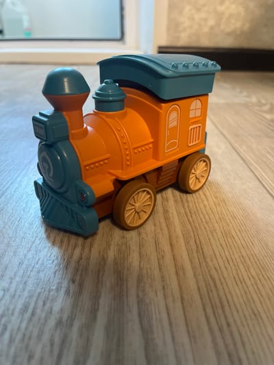 This image shows a close-up, angled view of an orange and teal toy train. The train appears to be made of plastic and is resting on a wooden surface with a visible grain pattern. The train has several distinct features, including cream-colored wheels with spokes, a teal-colored front with a rounded shape, and an orange body. The orange body has embossed details that resemble windows and a smokestack. There are also small, circular raised details on the side of the train. A teal-colored piece with a ridged texture is attached to the bottom rear of the train.
Visible text on the toy includes the word "TRAIN" in white capital letters on a black rectangle. There is also a circular emblem on the teal section with the letter "C" inside.
The lighting suggests an indoor setting, possibly a room with natural light. No people are visible in the image. The background is blurred, but it appears to be a wall or a door. The overall impression is of a child's toy photographed in a casual setting. The image is shot from a slightly overhead perspective, with the train oriented diagonally.