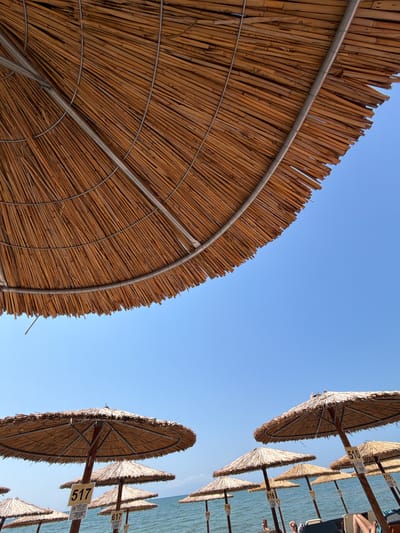 The image presents a low-angle view looking upwards at a collection of straw or thatch beach umbrellas against a clear blue sky, set in Alexandroupolis, Greece. The dominant subject is the underside of a large, circular beach umbrella in the foreground, showcasing its intricate woven texture and metal support framework.
Extending into the mid-ground and background on the left, numerous similar umbrellas line what appears to be a beach or coastline. Below these distant umbrellas, the calm, blue-green sea is visible, suggesting a clear and sunny day. Several umbrellas feature rectangular white tags with black numbers attached to their wooden poles, likely denoting specific spots or rental numbers. Clearly legible numbers include "517", "617", "817", "516", "616", and "716". Other partial numbers such as "715", "615", "618", and "816" are also discernible. A small, unreadable sign with symbols, possibly conveying beach rules, is affixed beneath the "517" tag.
Faint outlines of people, specifically legs and torsos, can be observed relaxing on sun loungers beneath some of the umbrellas on the left, indicating ongoing leisure activity. The bright, cloudless blue sky, occupying a significant portion of the frame, confirms a beautiful, sunny day. The scene captures a typical summer setting of relaxation at an organized beach.