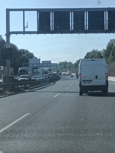 The image captures a highway scene in Kunice, Czechia, likely during the daytime given the bright sunlight casting shadows. The weather appears clear and sunny, contributing to good visibility. The primary subject is a traffic jam on a multi-lane highway. In the foreground, a white van is prominently featured, with its rear license plate clearly visible as "4SP 2408". The van is in the right lane, moving with the flow of traffic. Several other vehicles, including cars and trucks, are visible in the middle and left lanes, also stopped or moving slowly. A large overhead gantry with dark rectangular panels spans across the highway, possibly displaying traffic information or signs, though the details on the panels are indistinct. Beyond the highway, lush green trees line the roadside, indicating a rural or semi-rural setting. The overall mood is that of a typical, albeit congested, highway commute on a clear day.