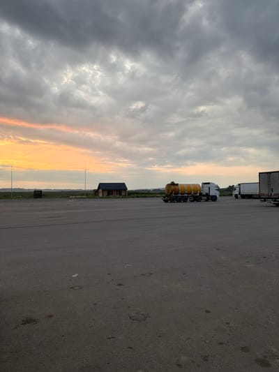The image depicts an outdoor scene at dusk, likely in a semi-rural or industrial area, possibly near Усть-Сыны, Russia, given the provided context. The foreground is dominated by a large, empty asphalt lot. In the middle ground, to the left, a small, wooden building with a dark roof is visible, flanked by tall poles. Further to the right, several semi-trailer trucks are parked. One truck in the center has a distinctive yellow cylindrical tank. The sky above is filled with dramatic, heavy clouds tinged with the orange and yellow hues of the setting sun, particularly on the left side of the frame. The overall atmosphere is overcast and quiet, with no people or visible activity other than the parked vehicles.