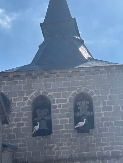 The image depicts the upper section of a stone church bell tower in El Espinar, Spain, against a clear blue sky with some white clouds. The tower is constructed from grey, rectangular stone blocks. A dark, possibly slate, conical roof with a spire tops the stone structure, with sunlight reflecting off its surfaces.
Below the roofline, two prominent arched openings are visible in the stone facade.
1. The leftmost fully visible archway contains a large, dark bell. Perched on the lower ledge of this opening, in front of the bell, is a static white stork figurine, characterized by a white body, black wingtips, and red-orange beak and legs.
2. The rightmost archway also contains a large, dark bell, with decorative vertical wooden slats positioned behind it. Another identical white stork figurine stands on the lower ledge of this opening, in front of its respective bell.
A third partial archway is visible to the far left, featuring a large, untidy bird's nest on its exterior left side. No human activity or movement is present in the frame.