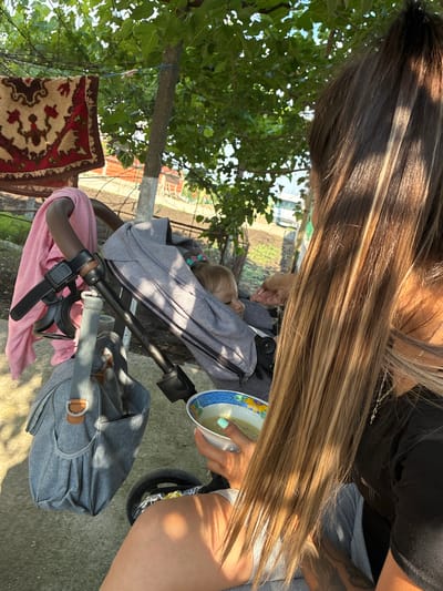 An overhead outdoor shot captures an adult and a child in Radievo, Bulgaria, during daylight hours. The adult, positioned to the right with long brown hair featuring blonde highlights cascading over their shoulder, wears a dark top and light-colored bottom, revealing a partial tattoo on the left arm. Their left hand, with light blue-painted fingernails, holds a white bowl with a blue and yellow floral pattern, containing a light-colored liquid. A silver necklace is visible on the adult's neck. A gray fabric stroller is positioned to the left of the adult. Inside the stroller, partially obscured by the adult's hair, a child with light-colored hair is visible, facing towards the left side of the frame, with one hand slightly raised. A pink cloth is draped over the left handle of the stroller, which also features a black accessory strap with a cup holder. A gray utility bag with a brown accent hangs from the stroller's frame. The ground beneath is concrete. The background features lush green tree foliage, with an ornate red and cream patterned rug hanging from a line on the left. A tree trunk with a white-painted base stands to the right of the rug. Distant structures, possibly houses or outbuildings, are vaguely visible beyond a dirt or field area. Sunlight casts strong shadows across the scene.