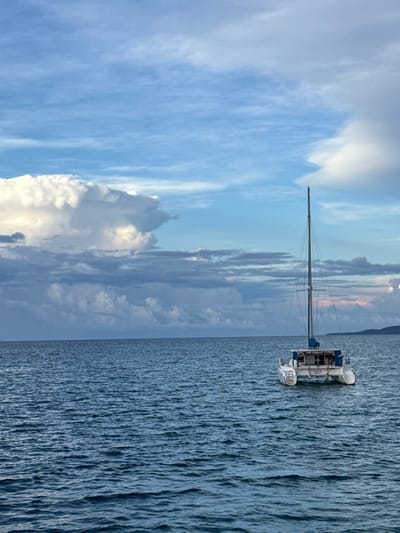 This image captures a marine environment in the city of Siquijor, Philippines. A white-hulled catamaran, equipped with a single central mast and blue accents on its superstructure, is stationary on the dark blue-green ocean, positioned with its stern partially visible towards the observer. The water surface displays minor ripples. The sky above is dominated by large, bright white cumulus clouds, intermingled with darker grey stratified clouds, and sections of lighter blue. A faint, dark landmass is discernible on the distant right horizon. No individuals, explicit actions, or direct interactions are present within the frame.
