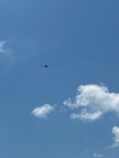 An aerial view captures a clear blue sky dotted with scattered white clouds. In the upper left quadrant of the image, a dark silhouette of a helicopter is visible, appearing to be in flight. The helicopter is small in the frame, suggesting it is at a significant altitude. No other objects or individuals are present in the scene, and the background is solely the sky and clouds. The location is identified as Velykyi Trostianets, Ukraine.