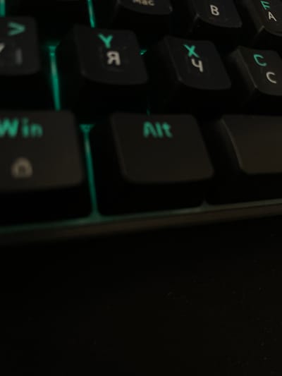 The image shows a close-up of a black keyboard with illuminated green keys. The visible keys include symbols and Cyrillic letters, suggesting a dual-language keyboard layout. Specifically, the keys labeled with "Win", "Alt", "Y", "Я", "X", "Ч", "C", and "C" are clearly visible. The perspective is angled downwards, focusing on the keys themselves. The background is dark and out of focus, indicating a low-light environment. The keyboard appears to be a standard computer keyboard, possibly with mechanical switches given the distinct keycaps. There is no indication of the city of Bornheim, Germany.