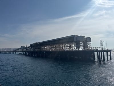 This image depicts a large industrial pier extending into a body of deep blue water under a bright, partially cloudy sky. The pier is a complex structure of metal scaffolding and large, elongated buildings, suggesting industrial operations such as loading or unloading. The architecture appears robust and functional, with visible cranes and conveyor belts. The sun is high in the sky, casting highlights on the water and indicating it is likely midday. The weather appears clear with some wispy clouds. The vast expanse of water and the industrial scale of the pier suggest a coastal or port location. No people are visible. The overall mood is one of industrial activity and vastness. There is no visible text in the image. The location is Mers El Hadjadj, Algeria.