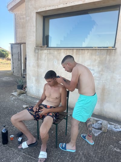 Two young men are outside a building, possibly in Ragusa, Italy, with one man sitting on a chair and the other standing. The man sitting is shirtless, wearing patterned swim trunks, and appears to be having his hair cut by the standing man. The standing man is also shirtless, wearing turquoise swim trunks, and is using clippers to cut the sitting man's hair. The environment appears to be a dry, somewhat arid outdoor space with a concrete ground and a textured, light-colored building wall in the background. A large window with a reflection of stairs is visible on the building. Several objects are scattered on the ground, including a large dark bottle, a small cup, a pair of flip-flops, and some plastic sheeting. The overall scene depicts a casual, DIY haircut taking place in an outdoor setting.
