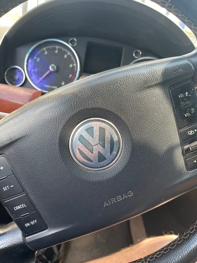 The image shows a close-up view of the steering wheel and dashboard of a car. The steering wheel is black leather with stitching and features several buttons. A prominent circular emblem with the Volkswagen logo is centered on the airbag cover. Text indicating "AIRBAG" is visible on the steering wheel. To the left of the steering wheel, parts of the car's instrument cluster are visible, including a speedometer with a red needle pointing towards a blue shaded area. Below the speedometer is a smaller circular gauge. The scene appears to be an interior shot of a vehicle, likely parked. The lighting suggests it could be daytime. There are no people visible in the image. The location context provided is Aqköl, Kazakhstan.
