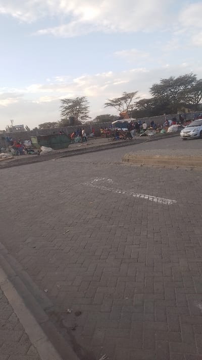 A paved road made of gray brick stretches into the distance, marked with a faded white arrow. Beyond the road, a bustling outdoor market thrives along a wall under sparse trees and a partly cloudy sky. People are gathered around stalls and carts, engaging in commerce. A green dumpster sits amidst the activity, and a silver car is parked nearby. The scene captures a busy day at a local market.