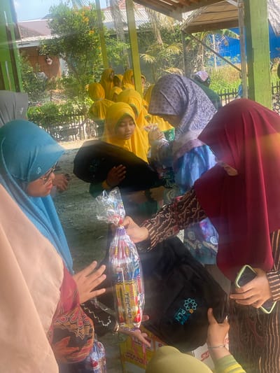 In Banyu Hirang, Indonesia, a group of young girls wearing yellow hijabs are gathered outdoors, possibly at a school or community event. The girls appear to be receiving gifts wrapped in plastic. In the foreground, three women, also wearing hijabs, are interacting with the children. One woman in a blue hijab is looking at a girl in a yellow hijab. Another woman, wearing a dark red hijab, is holding a gift bag and looking down. A third woman, wearing a purple patterned hijab, is also holding a gift. The background shows trees, a building, and a fence. The ground is covered with dirt. The overall scene suggests a charitable or celebratory event.
